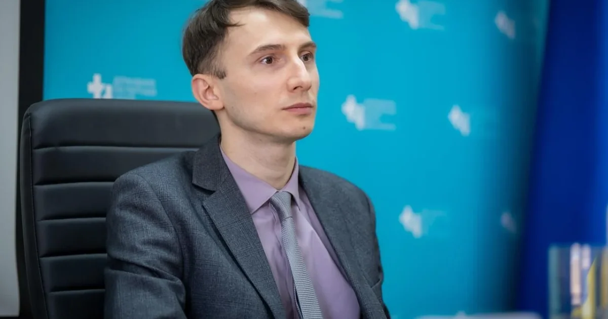 Чоловік у сірому костюмі та бузковій сорочці сидить у чорному кріслі на тлі синього банера