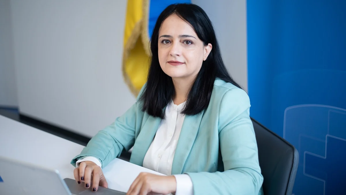 Жінка в бірюзовому піджаку сидить за столом із ноутбуком біля прапора